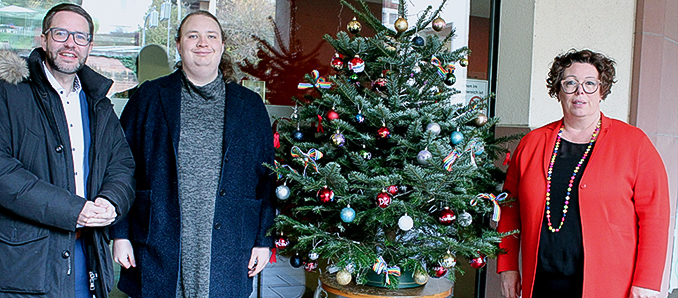 Freuen sich über den bunt geschmückten „Queeren Weihnachtsbaum“, der vor dem Main-Kinzig-Forum in Gelnhausen um Toleranz und Respekt wirbt und ein Zeichen gegen Hass setzt (von links) Landrat Thorsten Stolz, Selena Krieg (Verein Queer* Main-Kinzig und Erste Kreisbeigeordnete Susanne Simmler.