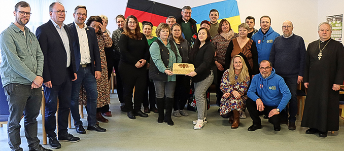 Die Caritas-Gäste in der Flüchtlingsunterkunft in Hilders-Steinbach mit den Flüchtlingen (und selbst gebackenen „Caritas-Kuchen“) sowie mit Vertretern von Politik und Kirche. Foto: Caritas